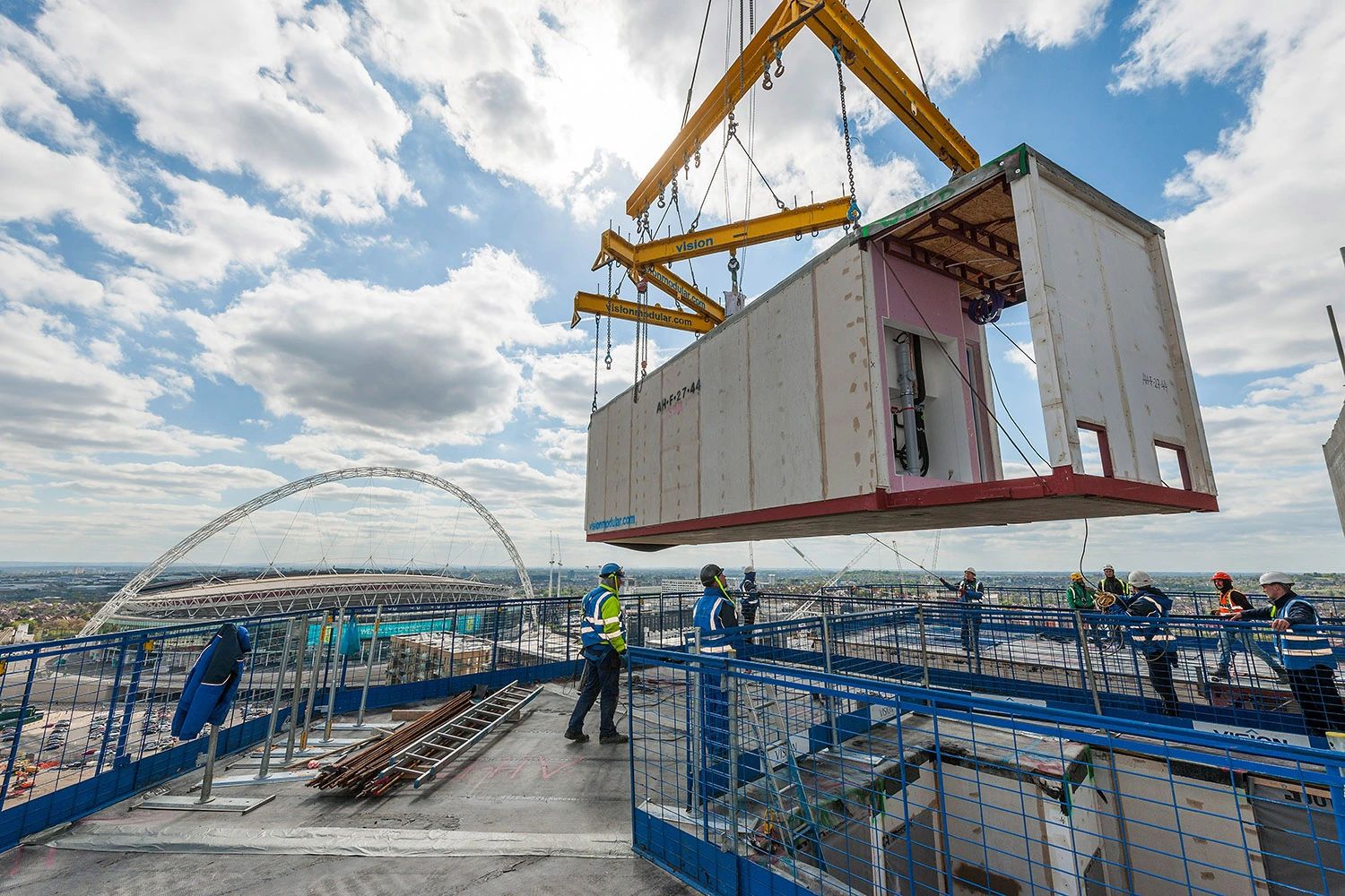 A modular building unit being lifted by crane on a construction site near Wembley Stadium.